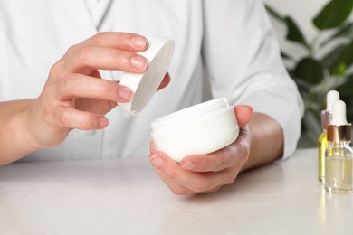 Photo of Dermatologist holding jar of cream at white table, closeup. Developing cosmetic product