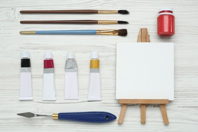 Photo of Easel with blank canvas, brushes and paints on white wooden table, flat lay. Space for text