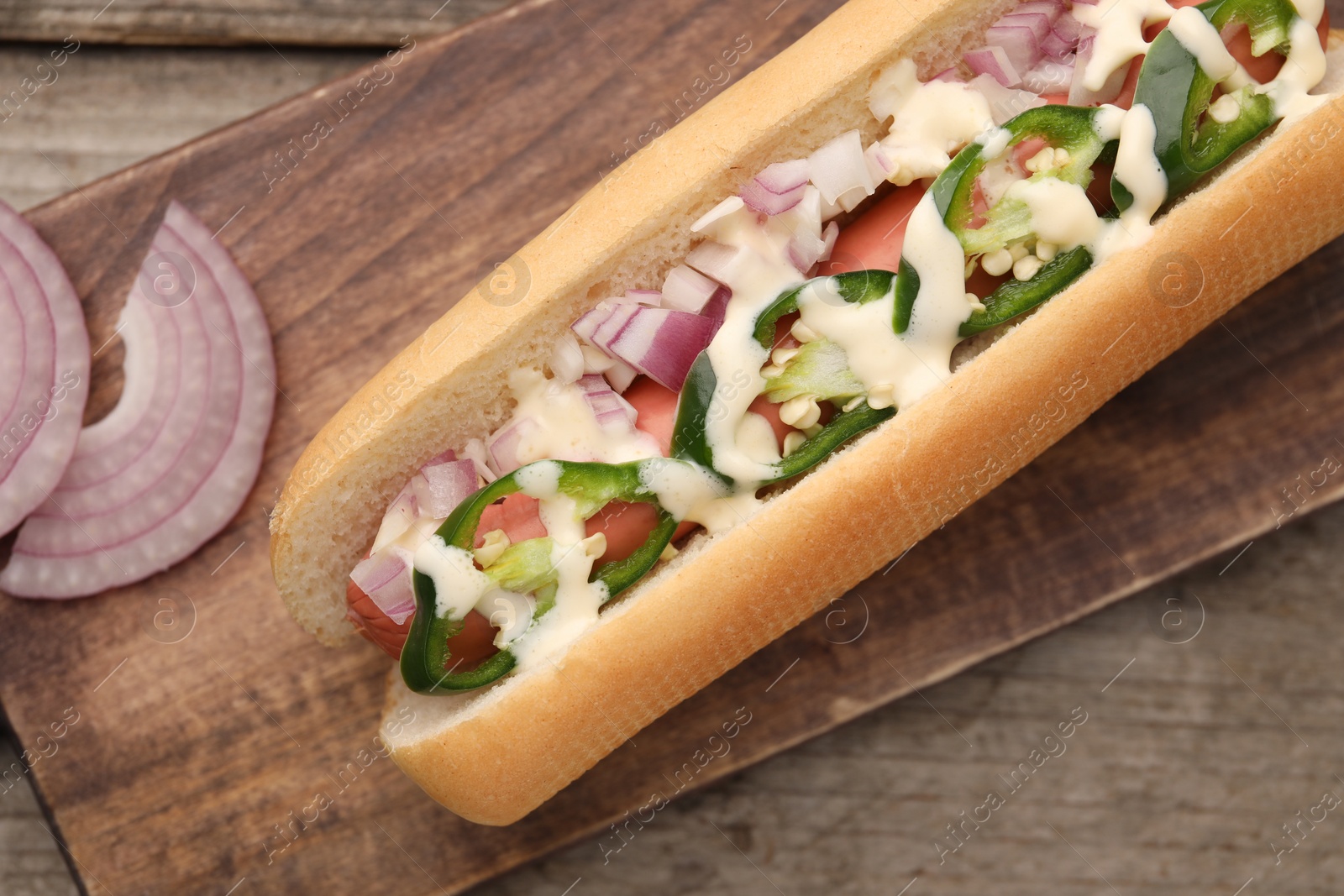 Photo of Delicious hot dog with onion, chili pepper and sauce on wooden table, top view