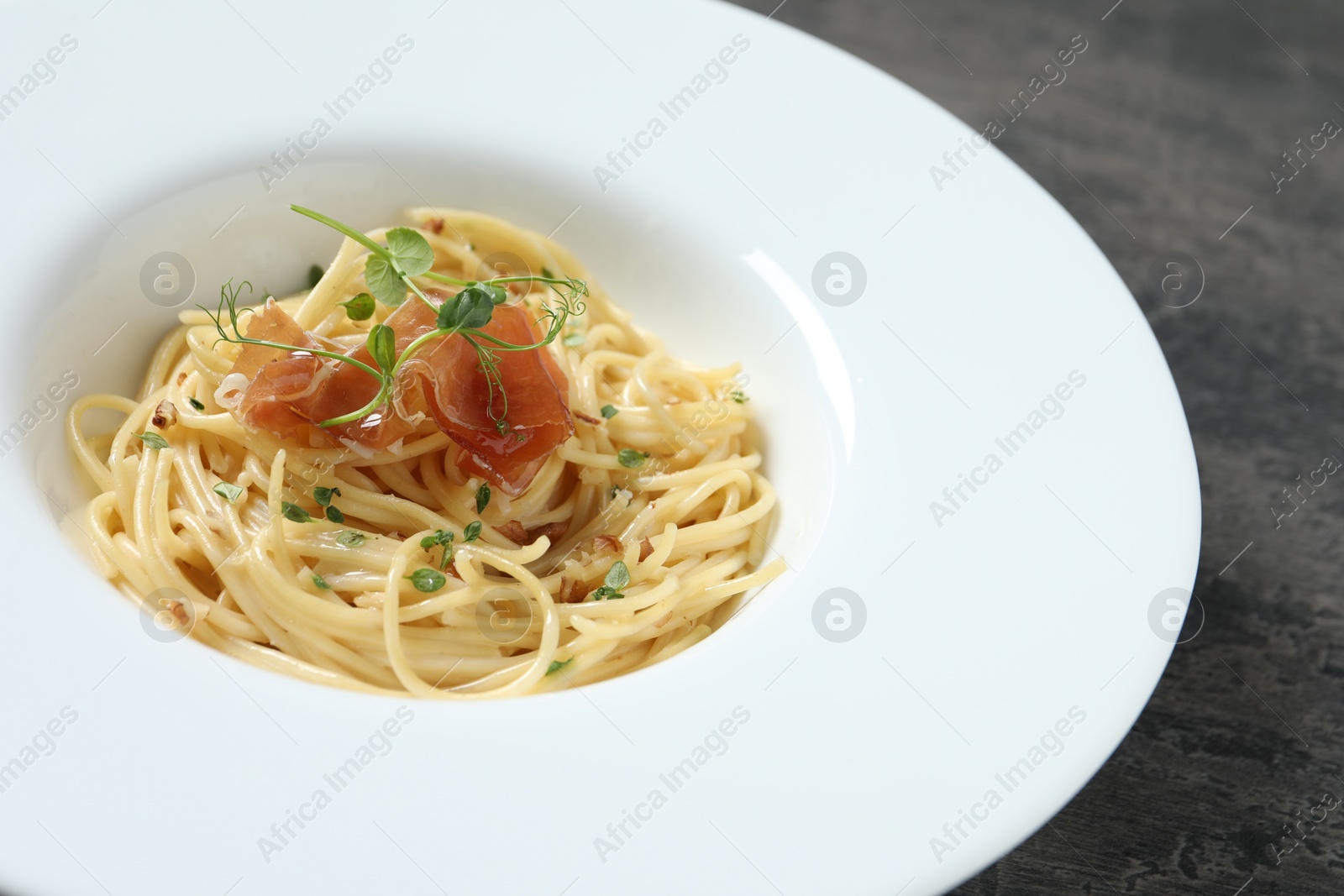 Photo of Tasty spaghetti with prosciutto and microgreens on grey table, closeup. Exquisite presentation of pasta dish