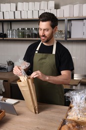 Photo of Happy seller putting pastry into paper bag at cashier desk in bakery shop