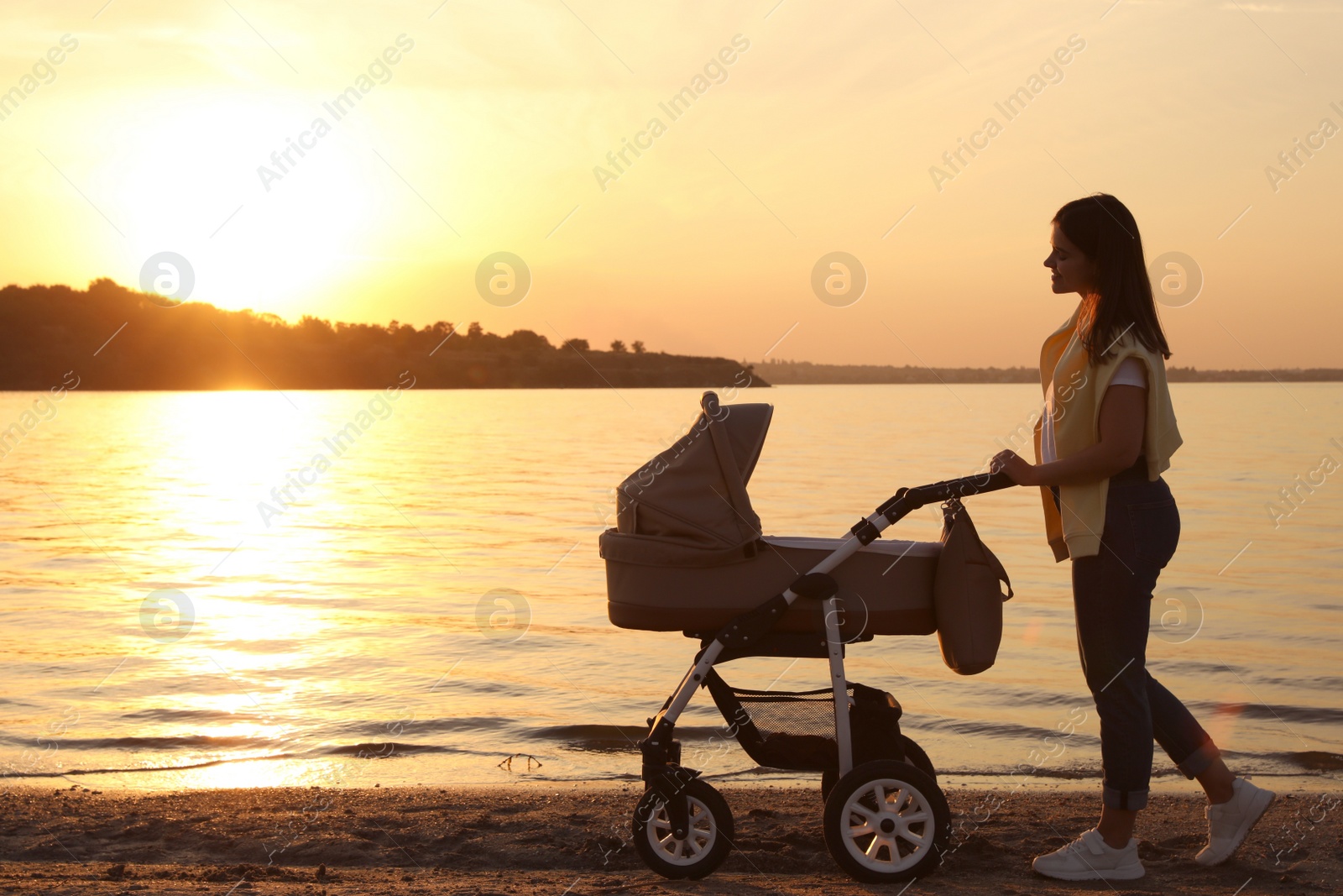 Photo of Happy mother with baby in stroller walking near river at sunset