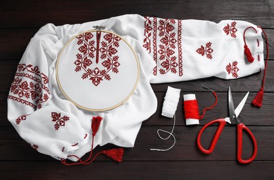 Photo of Shirt with red embroidery design in hoop, needle, scissors and threads on wooden table, flat lay. National Ukrainian clothes
