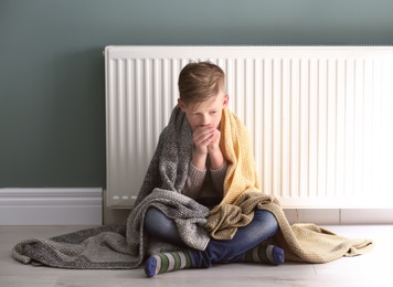Sad little boy suffering from cold on floor near radiator