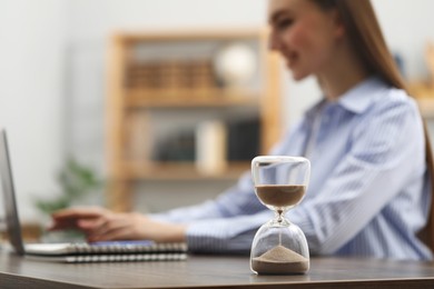 Photo of Hourglass with flowing sand on desk. Woman using laptop indoors, selective focus