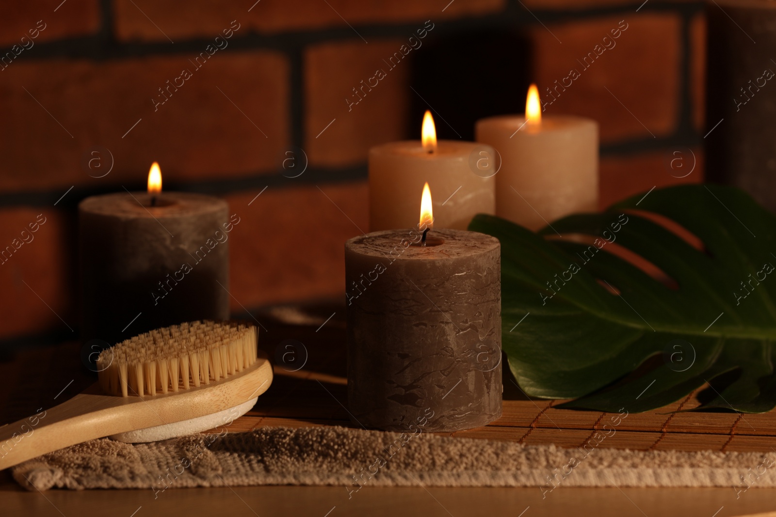 Photo of Spa composition with burning candles, massage brush and monstera leaf on wooden table near red brick wall