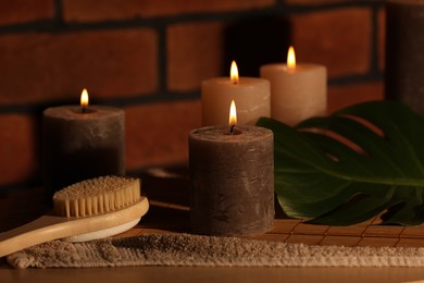 Spa composition with burning candles, massage brush and monstera leaf on wooden table near red brick wall