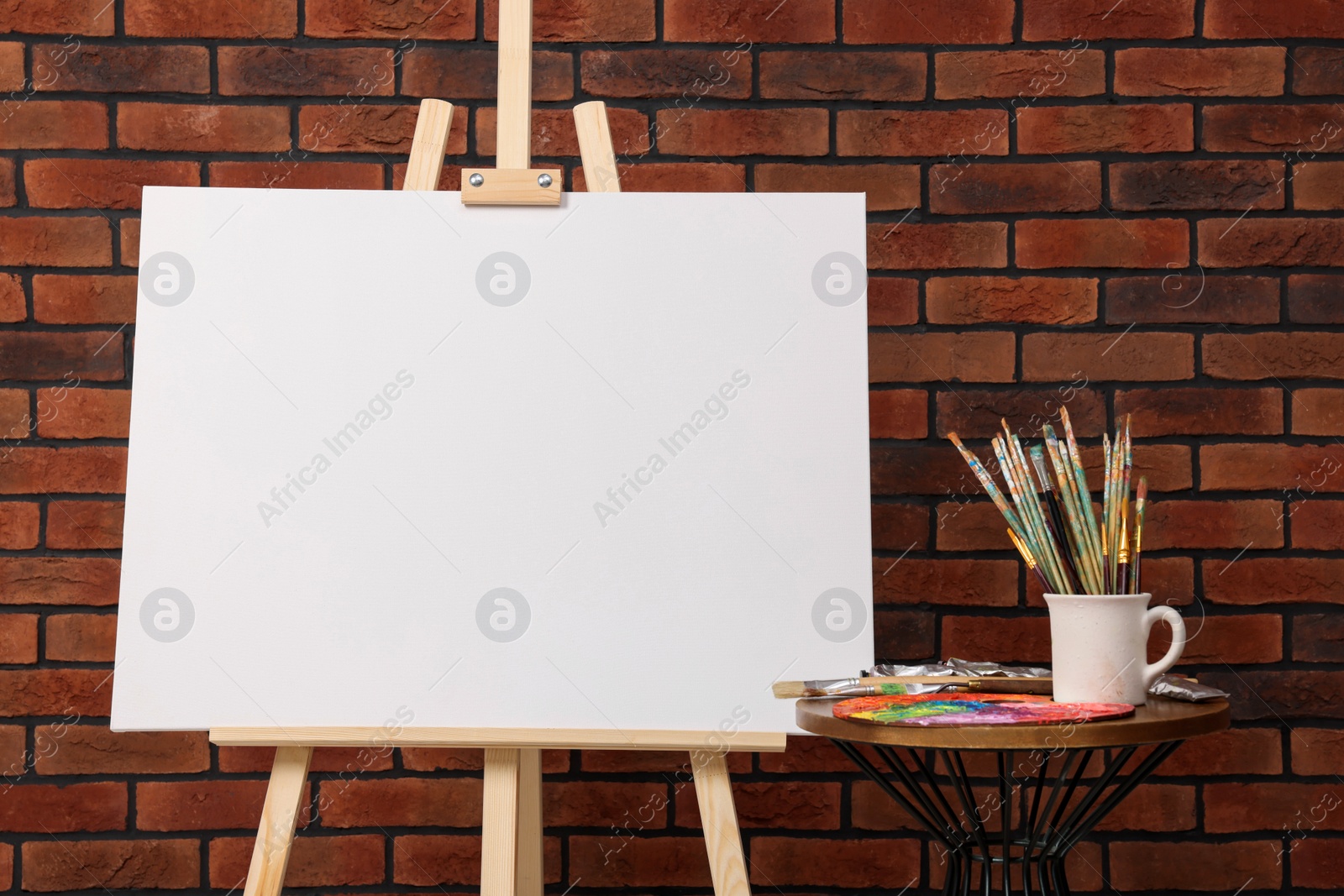 Photo of Wooden easel with blank canvas and different art supplies near brick wall, closeup