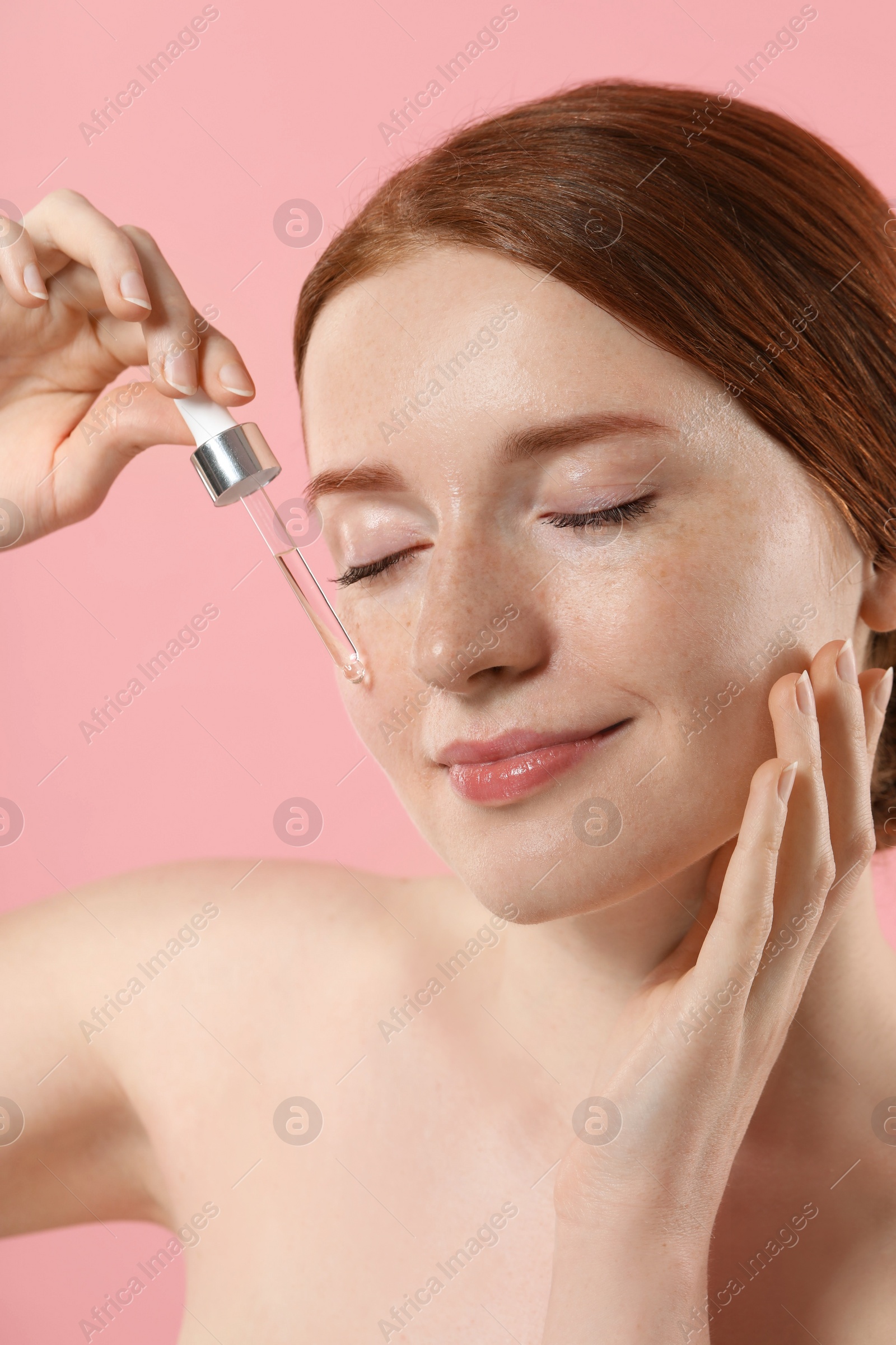 Photo of Beautiful woman with freckles applying cosmetic serum onto her face on pink background, closeup