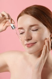 Beautiful woman with freckles applying cosmetic serum onto her face on pink background, closeup