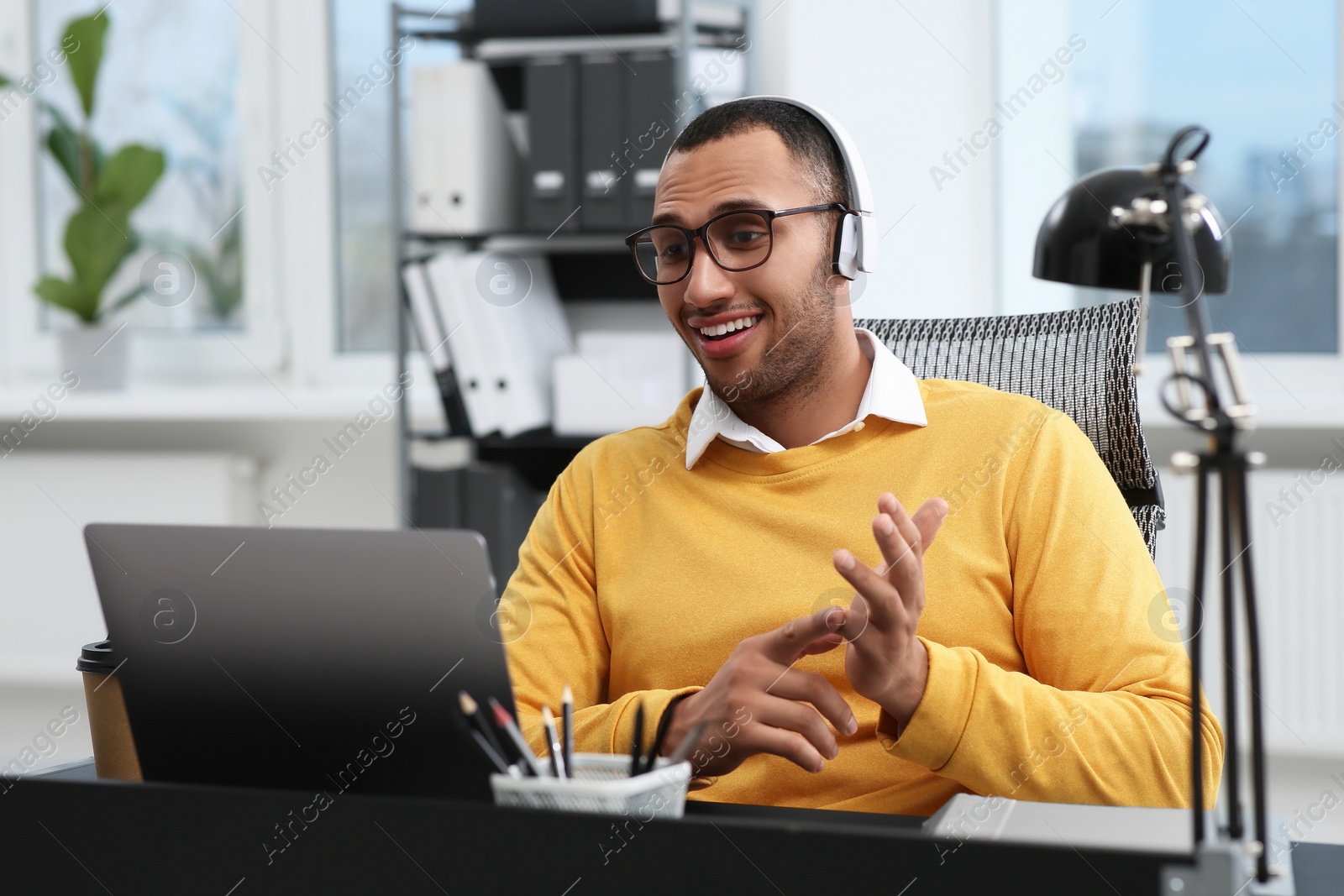 Photo of Young man with headphones having video chat via laptop at table in office