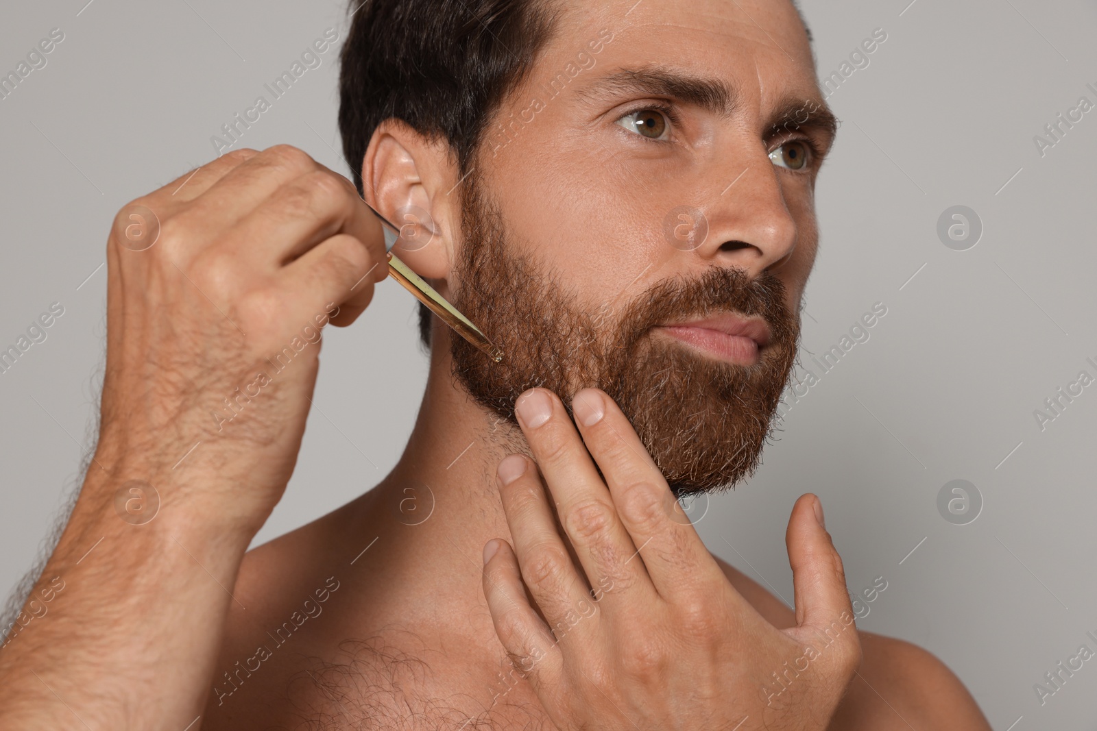 Photo of Man applying oil onto beard on grey background, closeup