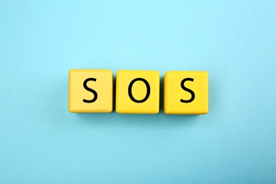 Photo of Abbreviation SOS (Save Our Souls) made of yellow cubes with letters on light blue background, top view