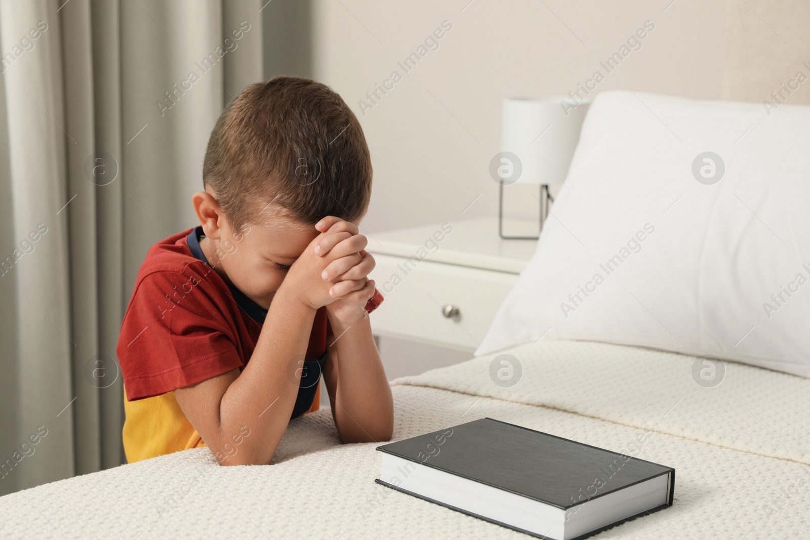 Photo of Cute little boy with hands clasped together saying bedtime prayer over Bible at home. Space for text