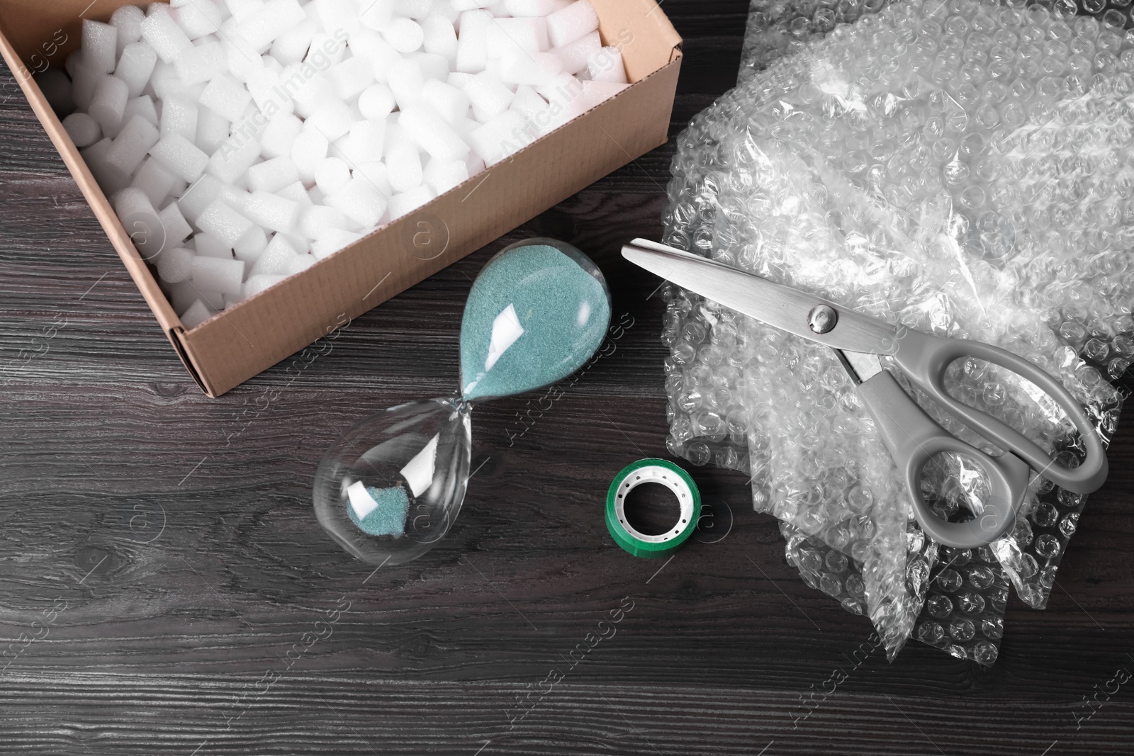 Photo of Sand hourglass, bubble wrap and packaging items on dark wooden table, above view