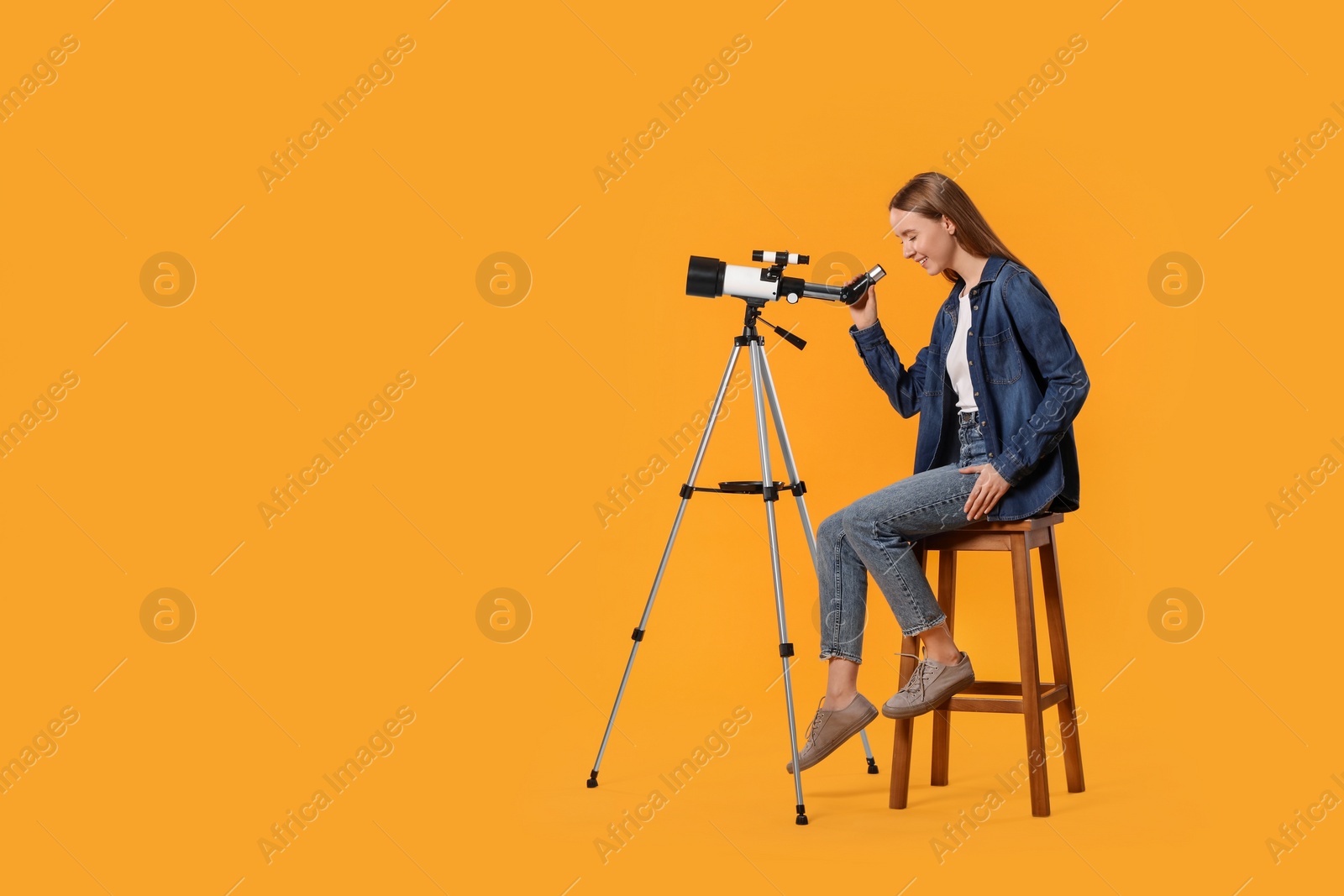 Photo of Young astronomer looking at stars through telescope on orange background, space for text