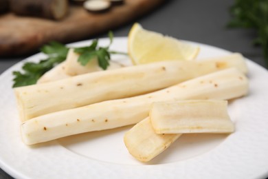 Photo of Cut raw salsify roots with parsley and lemon on white plate, closeup