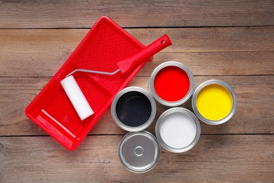 Photo of Cans of different paints, roller and tray on wooden table, flat lay