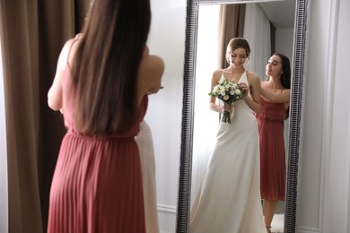 Gorgeous bride in beautiful wedding dress and her friend near mirror in room