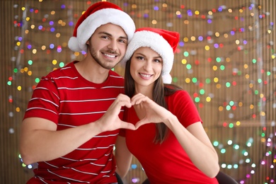 Photo of Young couple in Santa hats putting hands in shape of heart on blurred lights background. Christmas celebration