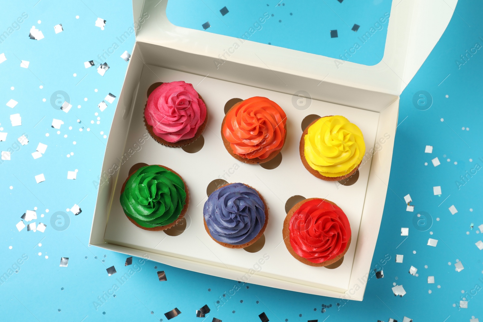 Photo of Box with delicious colorful cupcakes and confetti on light blue background, flat lay