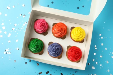 Box with delicious colorful cupcakes and confetti on light blue background, flat lay