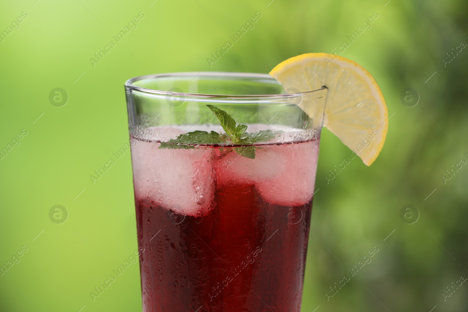 Photo of Refreshing hibiscus tea with ice cubes, mint and lemon in glass against blurred green background, closeup