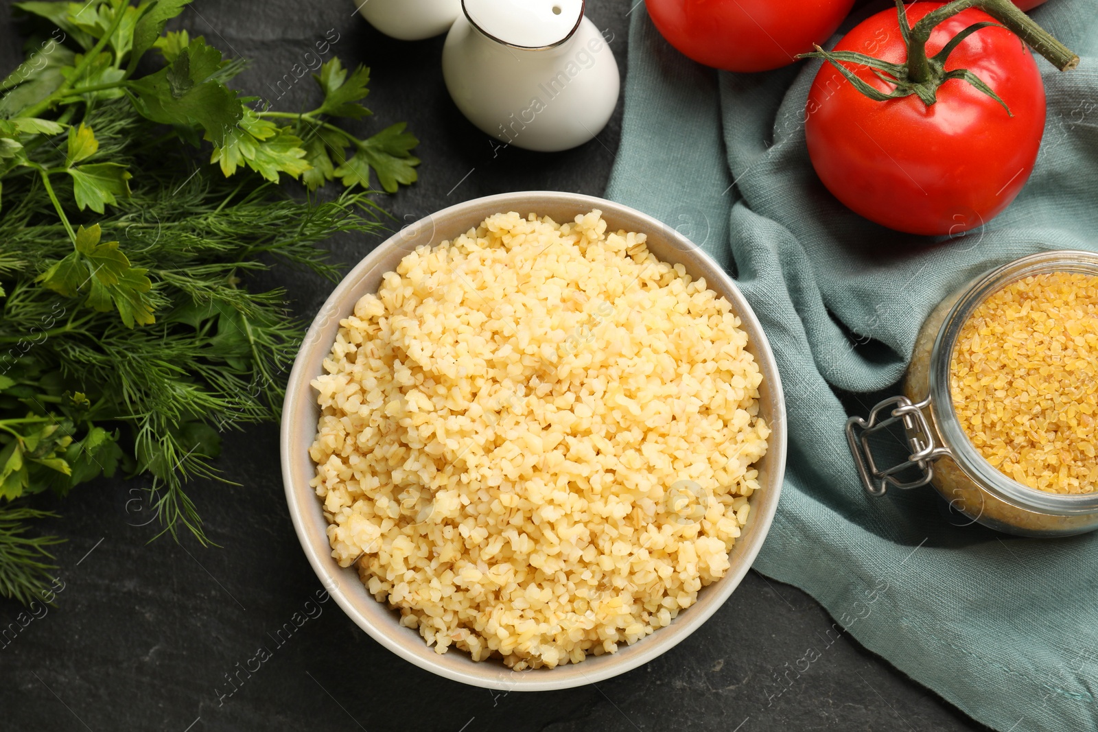 Photo of Delicious bulgur in bowl, dill, parsley and tomatoes on black table, flat lay