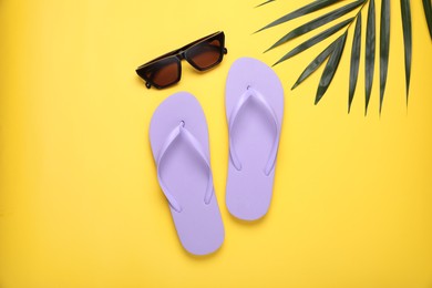 Photo of Stylish light purple flip flops, sunglasses and palm leaf on yellow background, flat lay