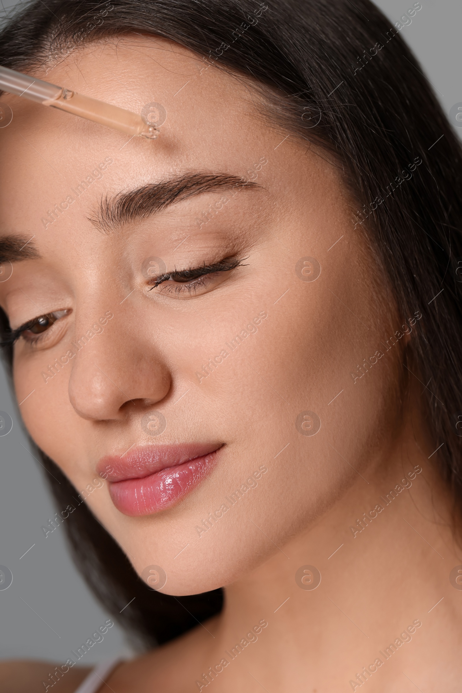 Photo of Beautiful young woman applying essential oil onto face on grey background, closeup