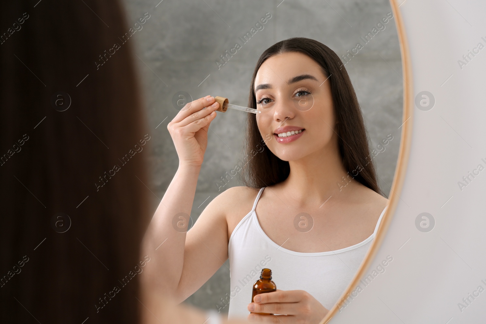 Photo of Woman applying essential oil onto face near mirror