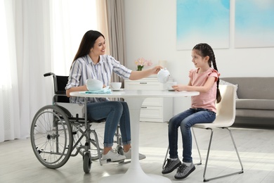 Mother in wheelchair and her daughter drinking tea at table indoors