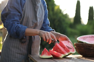 Photo of Woman cutting tasty ripe watermelon at wooden table outdoors, closeup