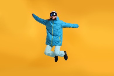 Winter sports. Woman with snowboard goggles jumping on orange background
