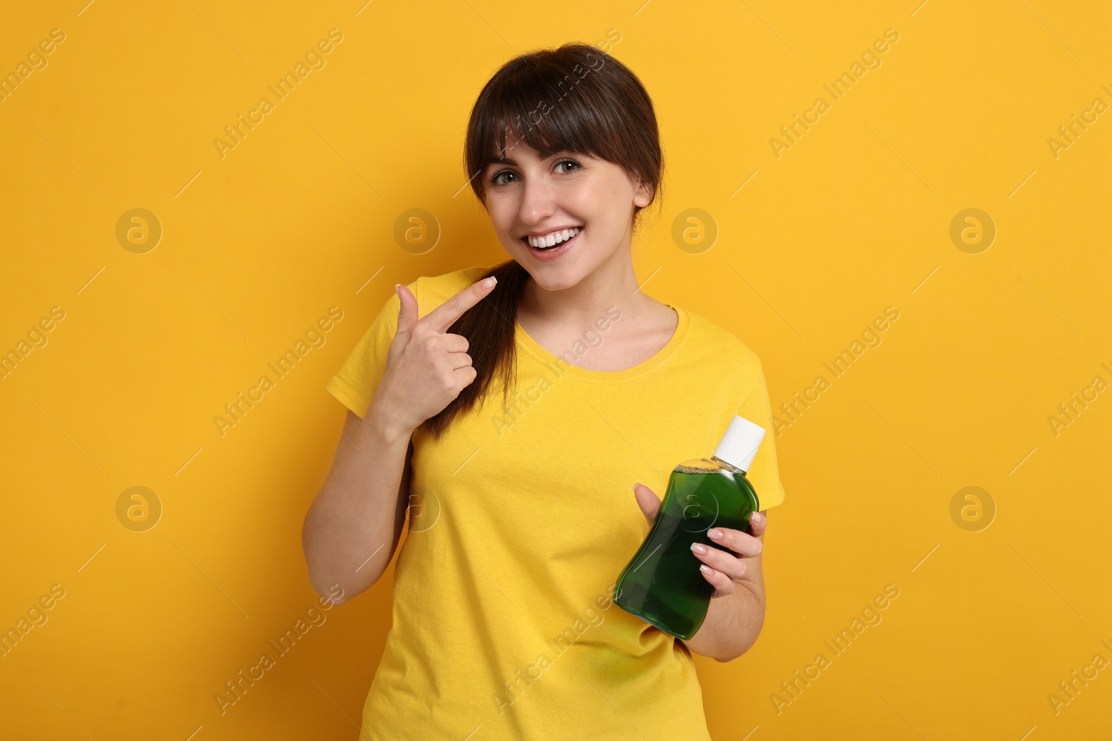 Photo of Young woman with mouthwash pointing at her healthy teeth on yellow background