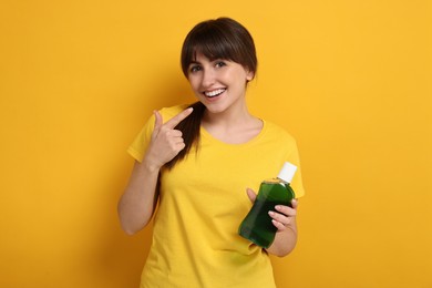 Young woman with mouthwash pointing at her healthy teeth on yellow background