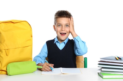 Emotional little boy in uniform with school stationery at desk against white background