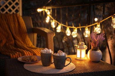 Photo of Cups with tasty cocoa and marshmallows on rattan table at balcony in evening