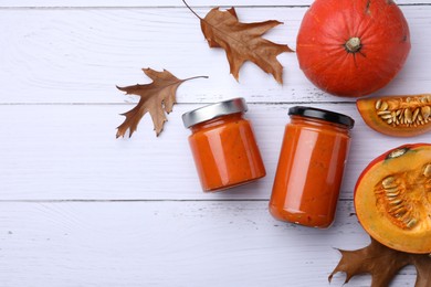 Photo of Jars of delicious pumpkin jam, fresh pumpkins and dry leaves on white wooden table, flat lay. Space for text