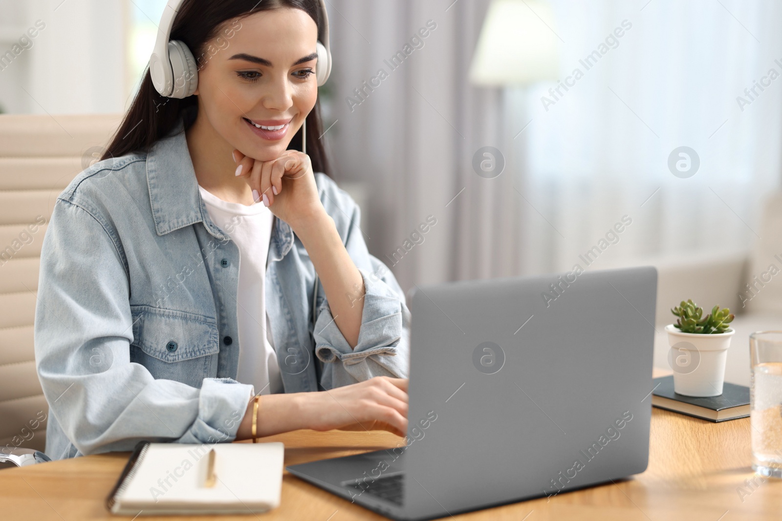 Photo of Young woman in headphones watching webinar at table in room