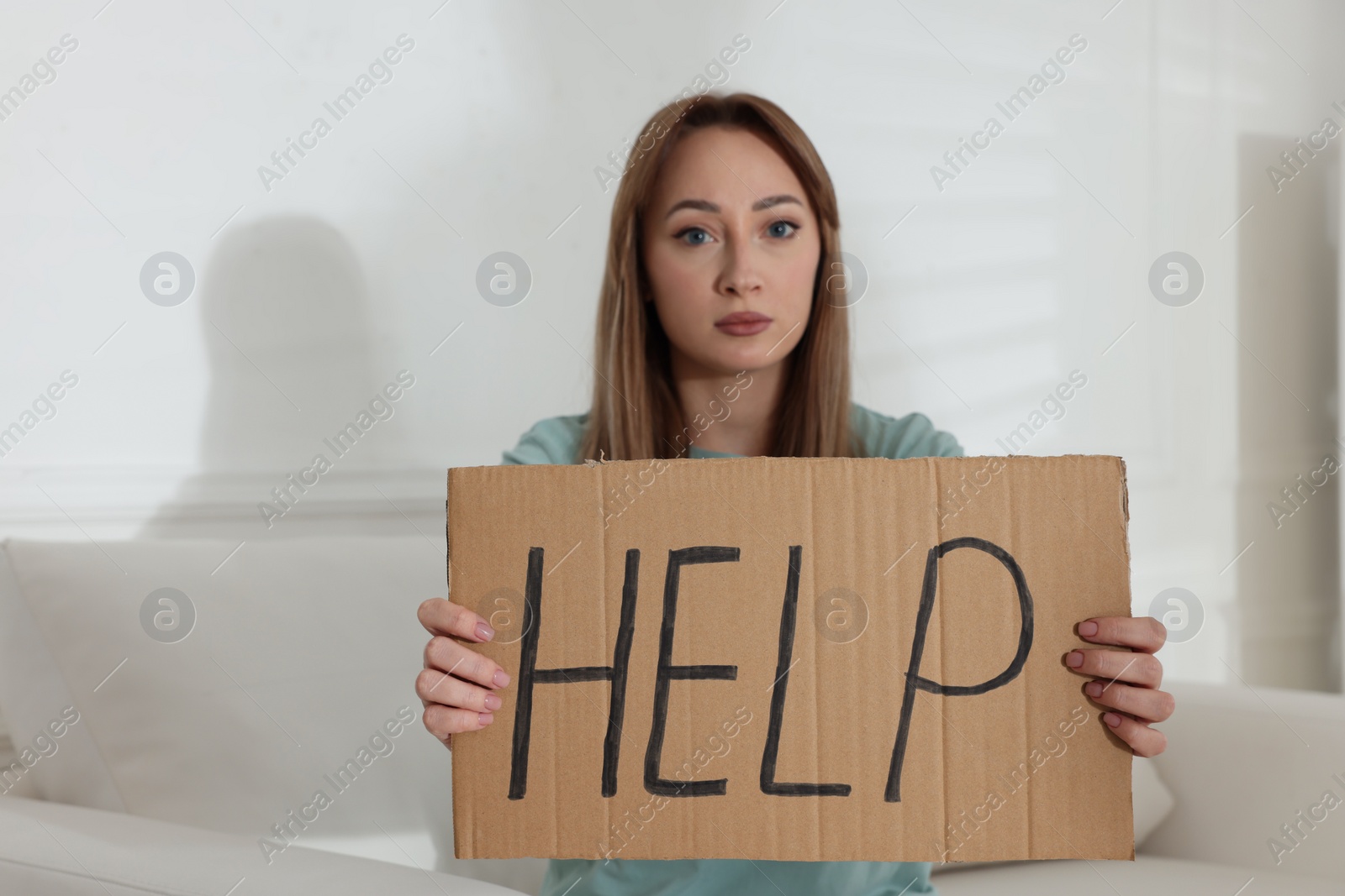 Photo of Unhappy young woman with HELP sign on sofa indoors