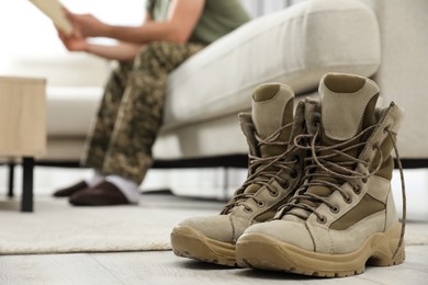 Photo of Soldier with magazine on sofa in living room, focus on pair of combat boots. Military service