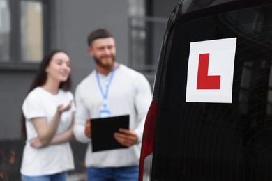 Photo of Learner driver and instructor with clipboard near car outdoors, selective focus on L-plate. Driving school
