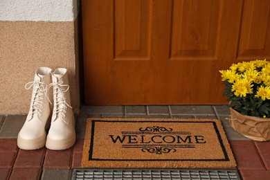 Photo of Door mat with word Welcome, stylish boots and beautiful flowers near entrance