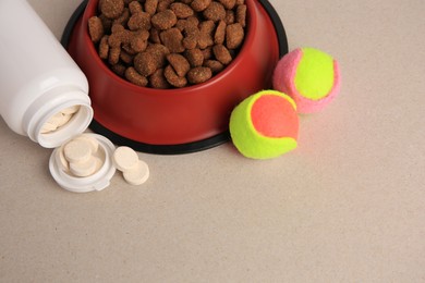 Bottle with vitamins, dry pet food in bowl and toy balls on beige background, space for text