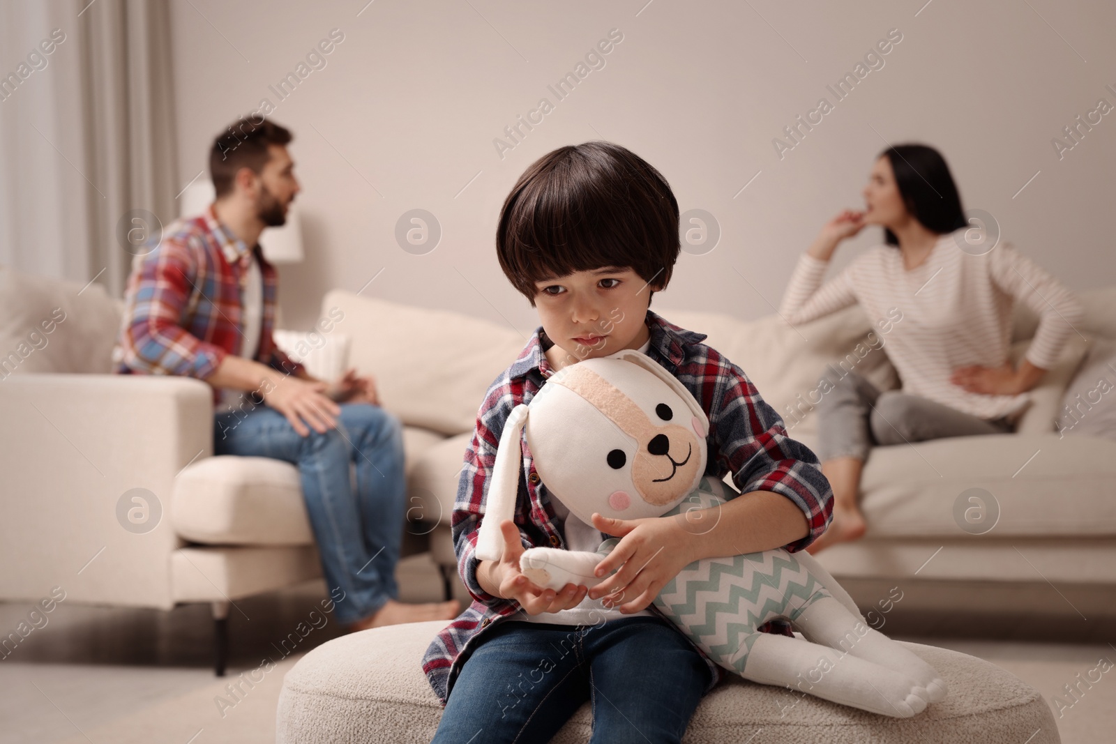 Photo of Sad little boy with toy and his arguing parents on sofa in living room