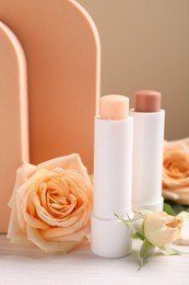 Stylish presentation of different lip balms with rose flowers on white table, closeup
