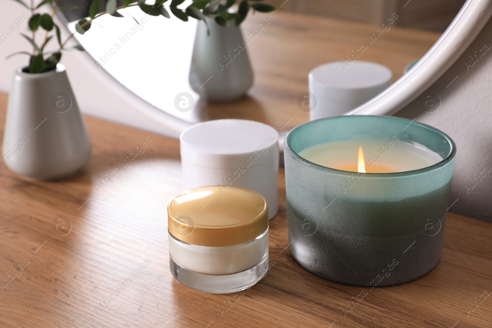 Photo of Burning candle, cosmetic products and vase on wooden dressing table, closeup