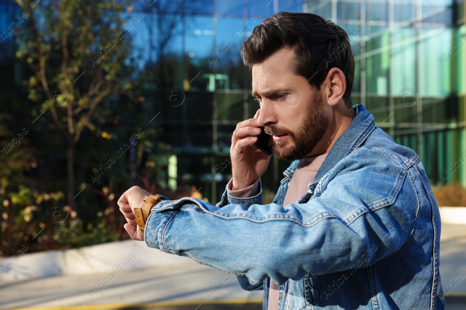 Photo of Emotional man talking on smartphone and looking at watch near building. Being late concept