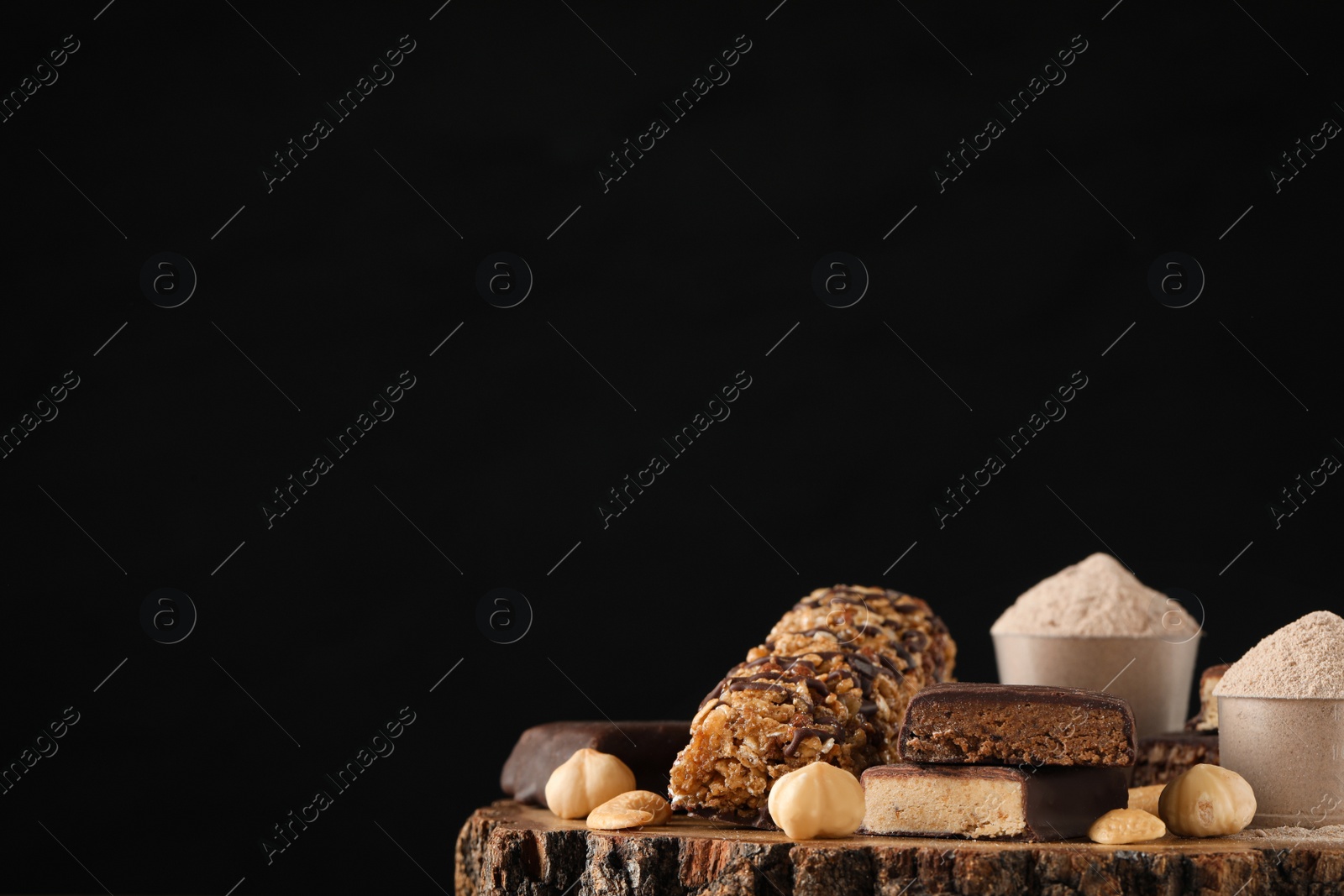Photo of Different tasty energy bars, nuts and protein powder on wooden stump against black background, space for text